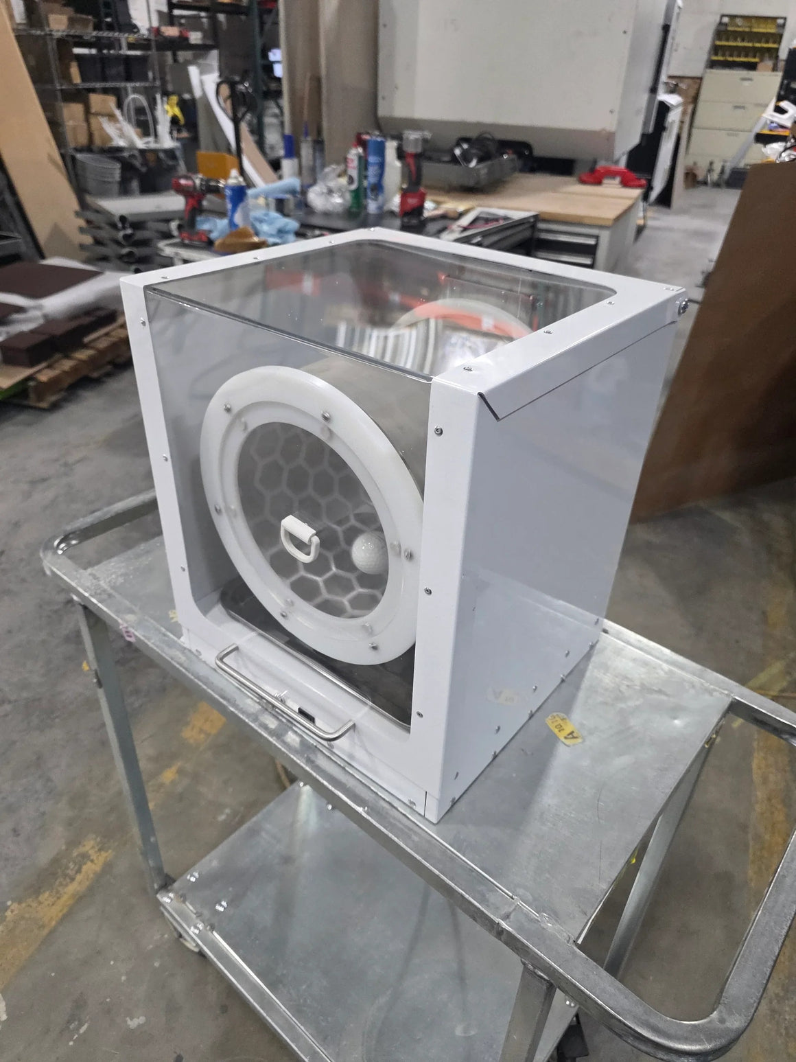 A white, clear-top extraction tumbler with a white circular grate on a silver metal cart in a workshop.
