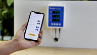 Hand holding iPhone showing Guardian device settings app; Blue and white monitor on wall.