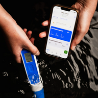 Hands hold a blue and white pH meter and a smartphone displaying water quality data, above dark, rippling water.