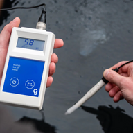 A hand holds a Bluelab soil pH meter, displaying a reading of 5.8, with the probe in dark water.
