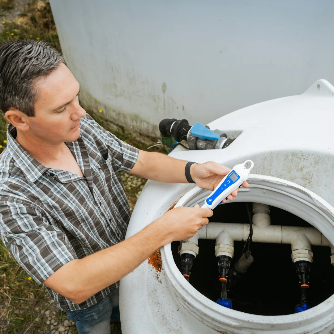 Man in plaid shirt uses a Bluelab pH PenPlus to test water in a white tank.
