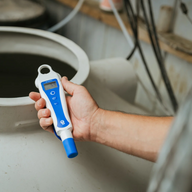 Blue Bluelab pH Pen Plus with 5.9 reading held in hand over a large white tank of murky liquid.