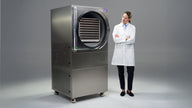 A woman in a lab coat stands next to a large stainless steel freeze dryer with its door open.