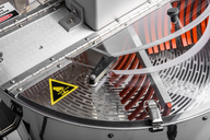 Close-up of a commercial bud trimmer's interior: clear plastic lid, metal frame, safety labels, and rotating blades.