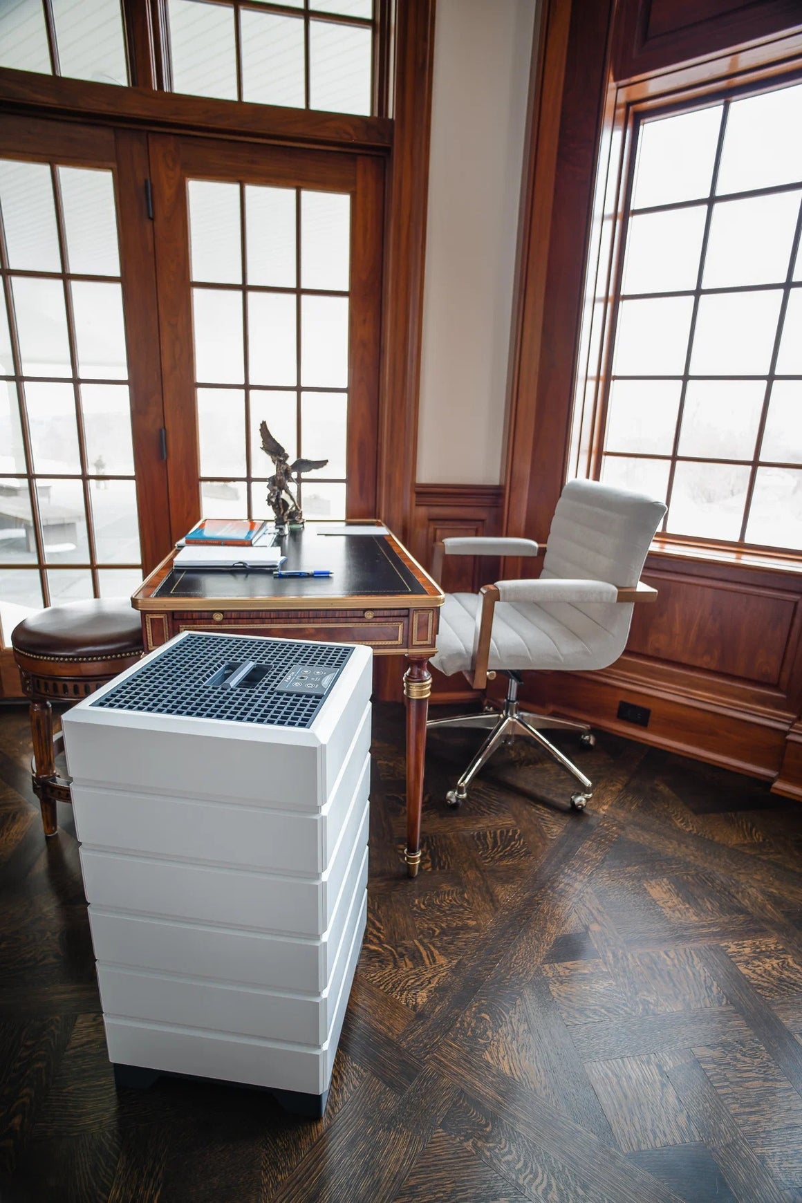An air purifier sits on a dark wood floor next to a desk and chair in a room with large windows.