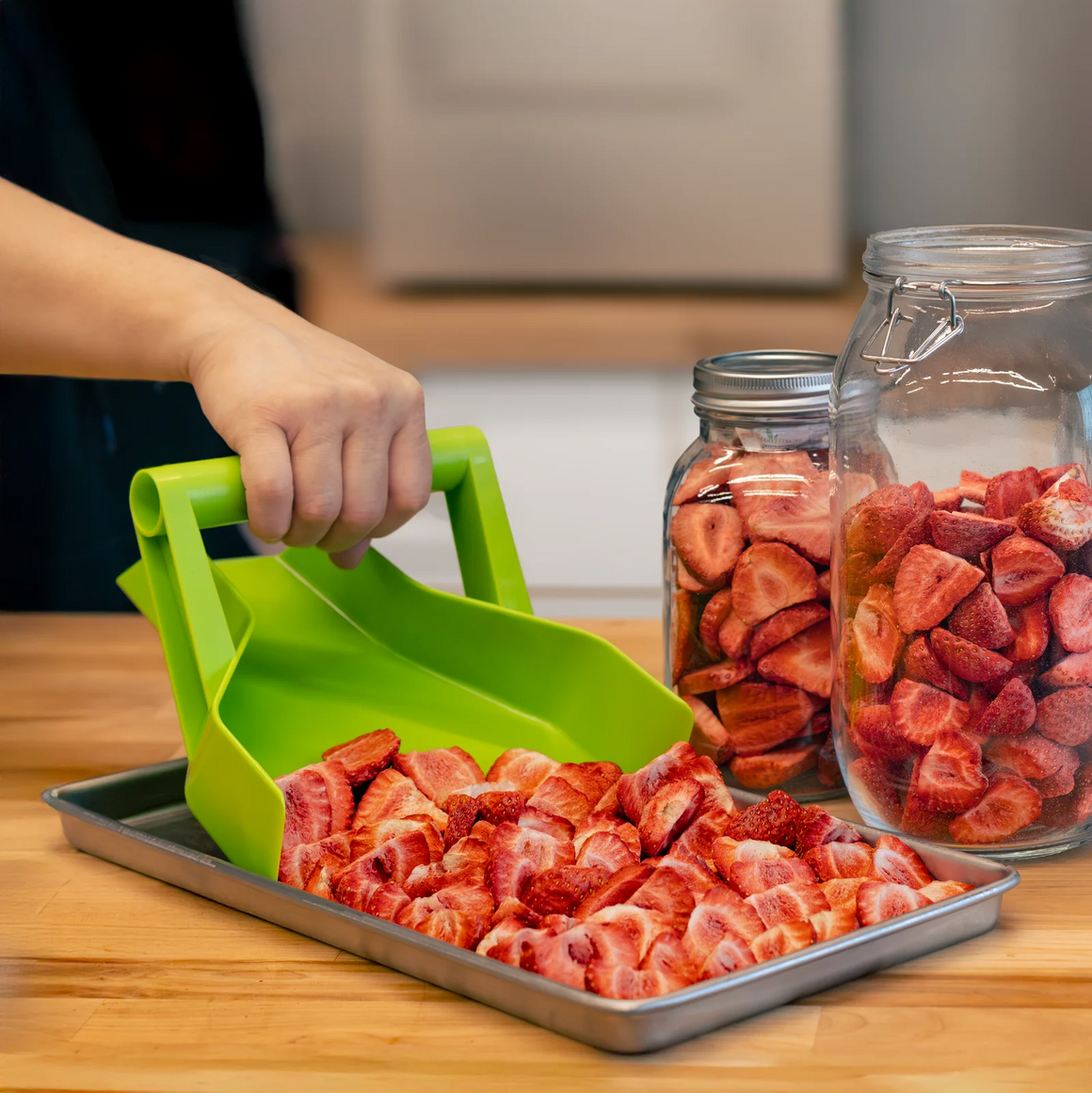 A person uses a green scoop to transfer sliced strawberries to a metal tray.