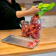 A person fills a jar with sliced strawberries using a green scooper.