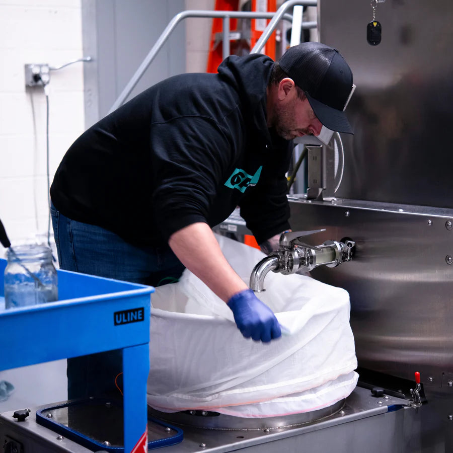 Man in black hoodie and blue gloves operates stainless steel equipment, collecting liquid in a large white bag.