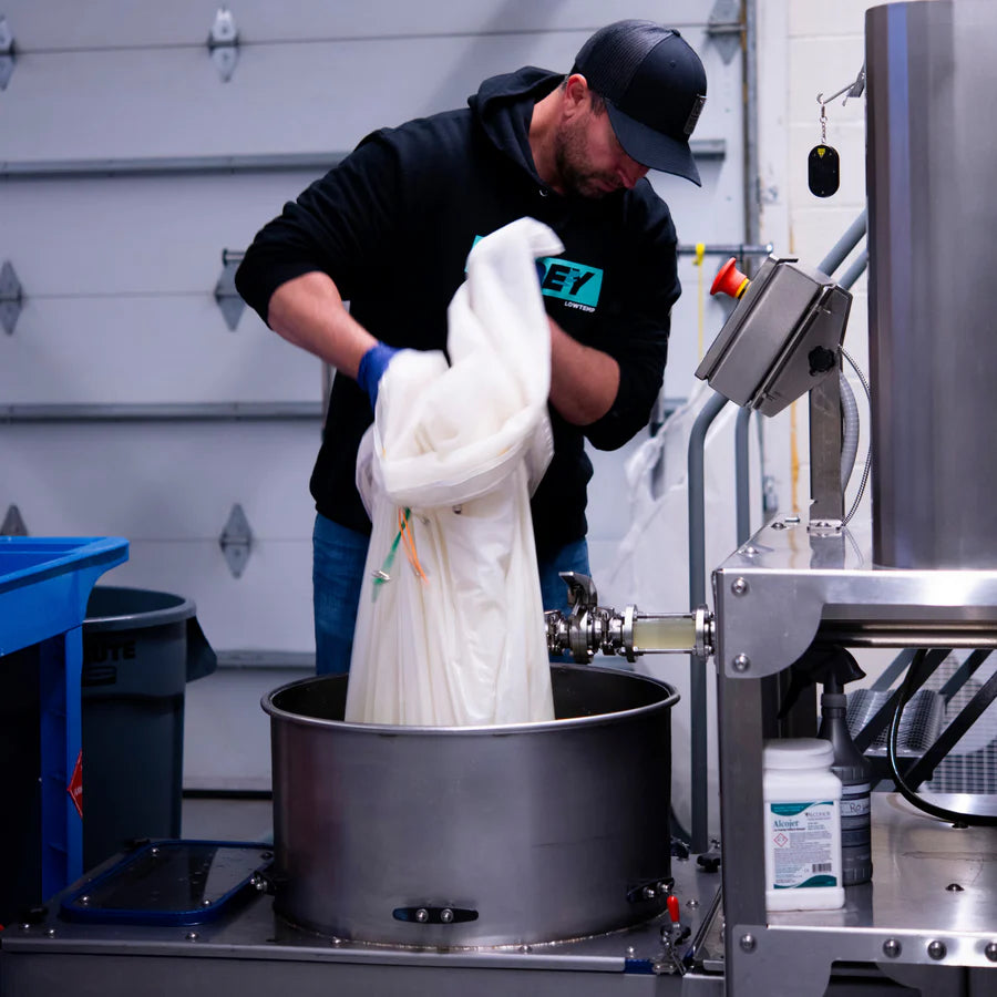 Man in black hoodie and blue gloves pours white material into stainless steel tank at a facility.