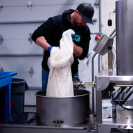 Man in black hoodie and blue gloves pours white material into stainless steel tank at a facility.
