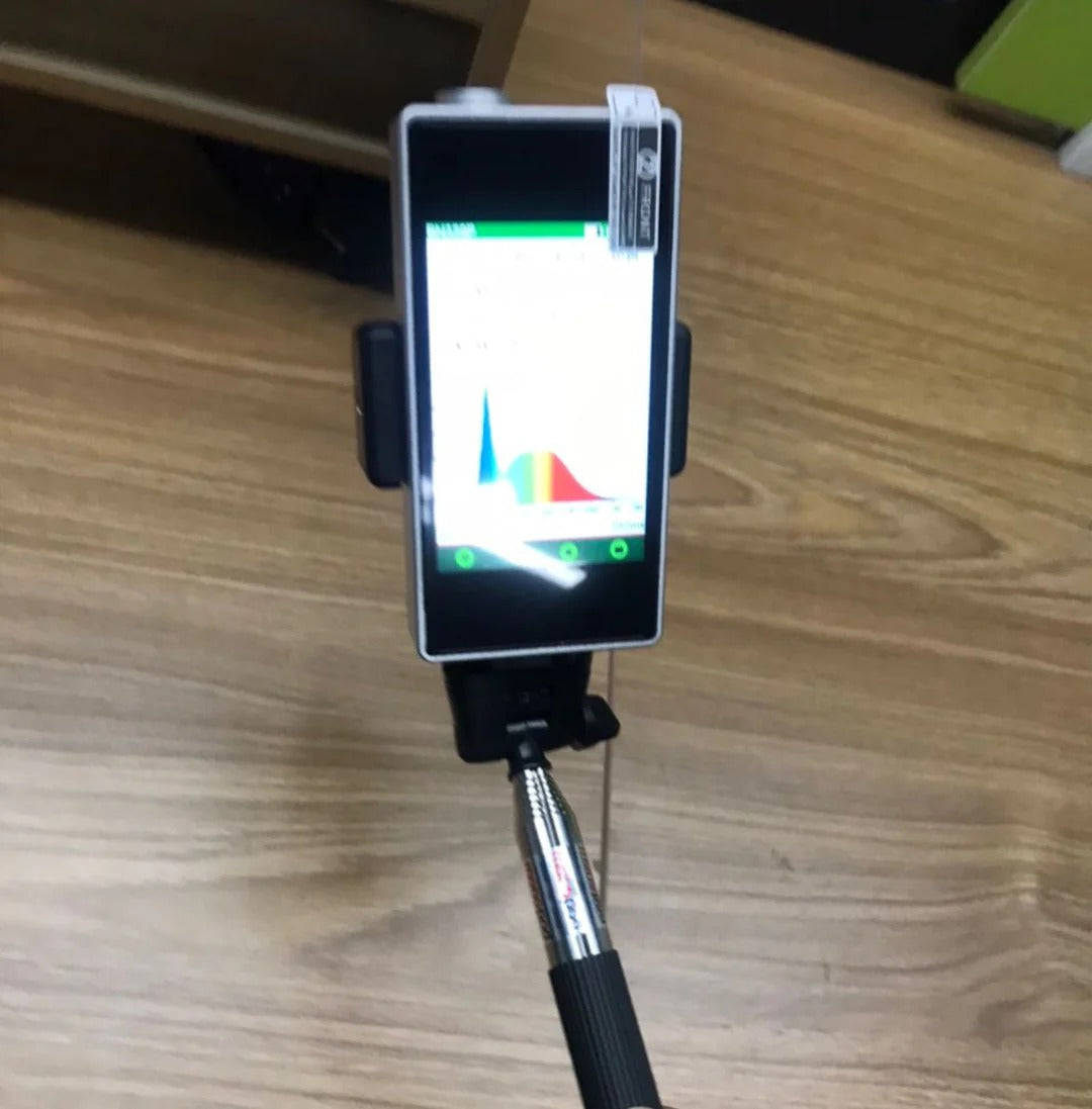 Smartphone with light spectrum graph on screen, held by selfie stick, over wood floor.
