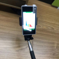 Smartphone with light spectrum graph on screen, held by selfie stick, over wood floor.