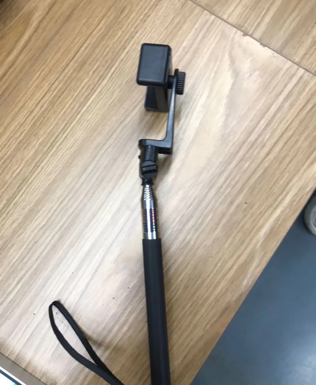 A black smartphone holder on an extendable selfie stick lies on a wooden surface.