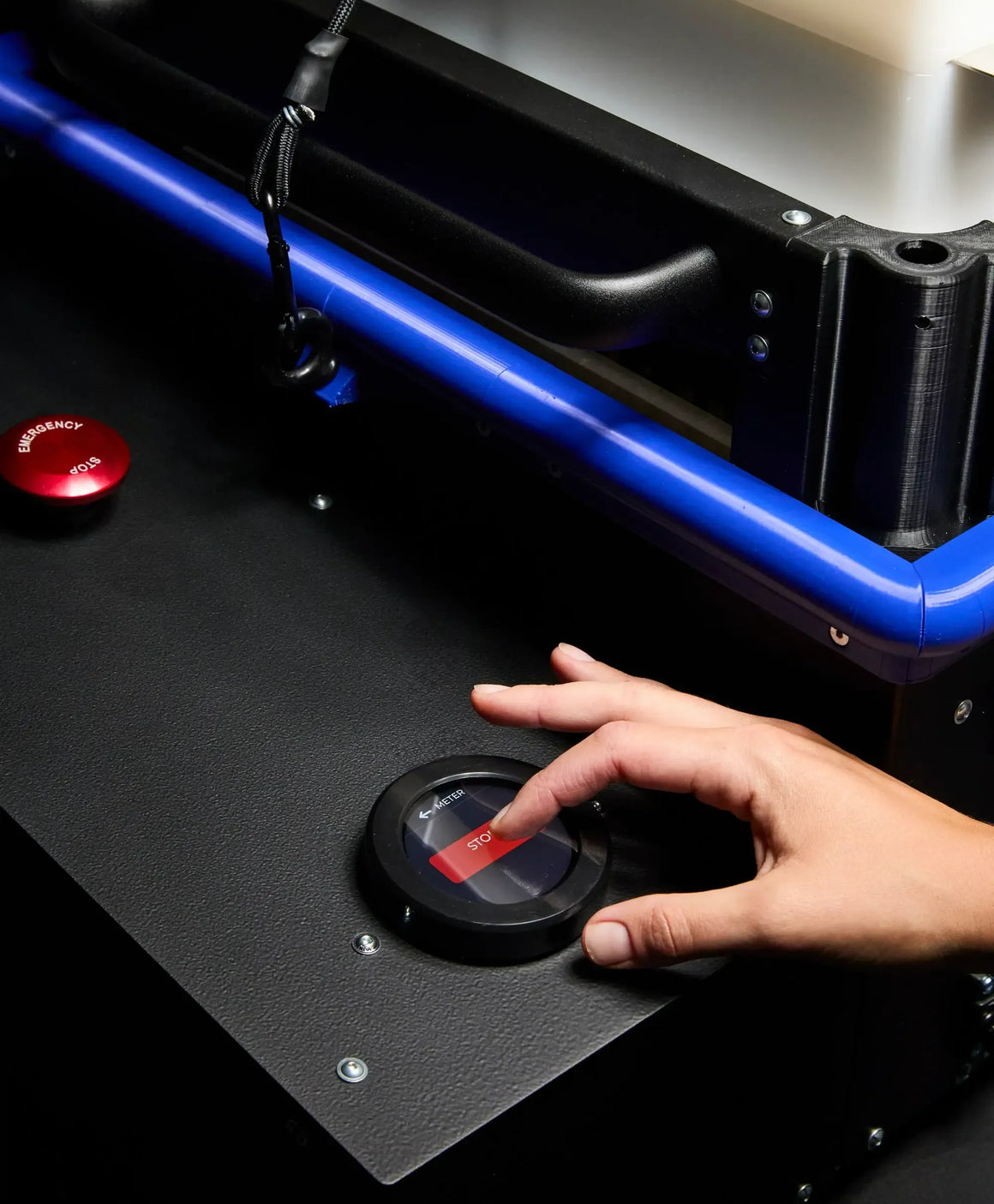 A hand presses a red "STOP" button on a black control panel with a blue railing.