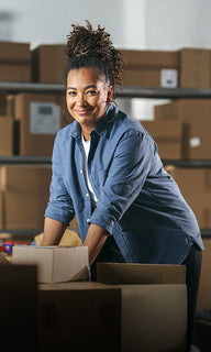 Woman with braided hair in a blue shirt, smiling and leaning over boxes in a warehouse.