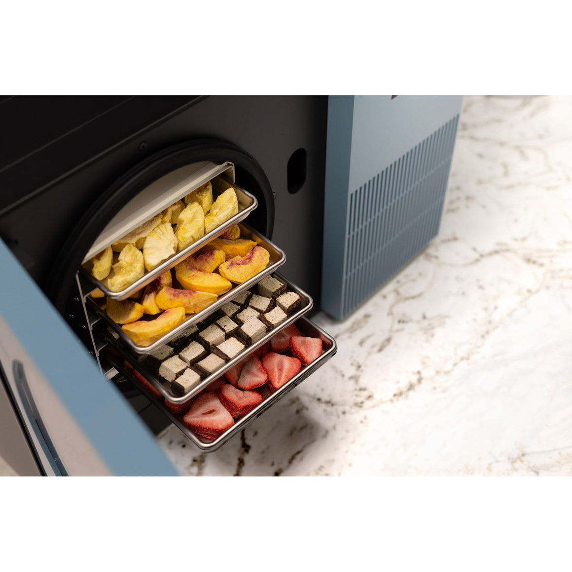 Pale blue Countertop 360 with the door open and trays of peaches, pineapple, chocolate pieces and strawberries pulled out on a marble counter