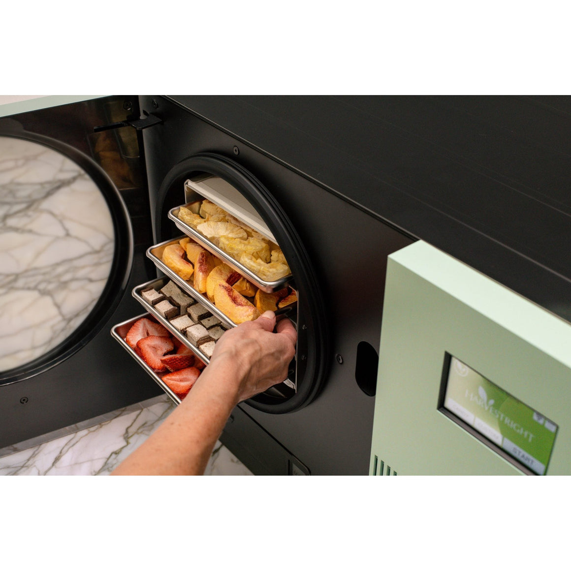 Hand sliding out trays of peaches, chocolate pieces and strawberries from the sage green Countertop 360 on a marble counter