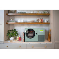 Sage green Countertop 360 freeze dryer on a kitchen counter under wooden shelves with a plant and jars of freeze-dried food