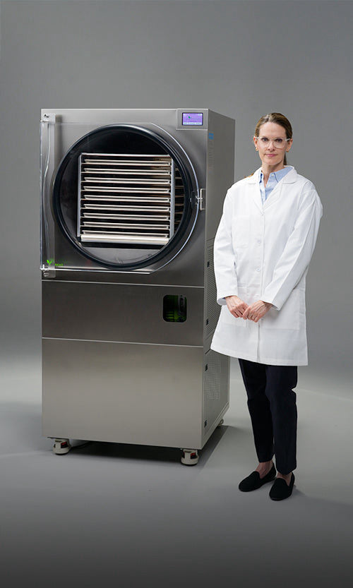 Scientist in lab coat stands next to a freeze-drying machine with trays inside.