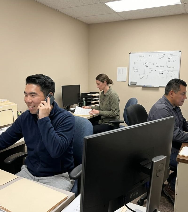 Three people working in an office: man on phone, woman with papers, man at computer.